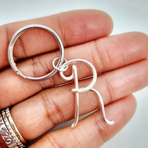 May include: A silver-colored keychain featuring the letter "R". The letter is crafted in a cursive style and attached to a round key ring. The keychain is held in a hand, showcasing the bright, reflective metal.