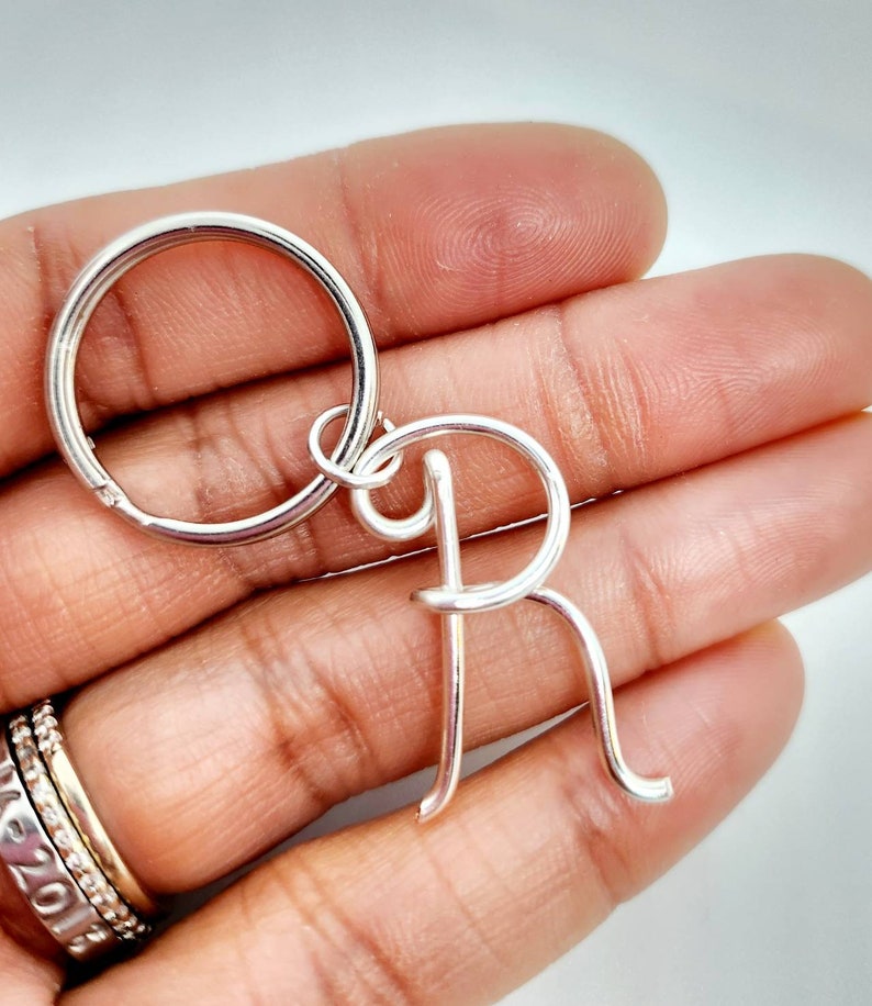 May include: A silver-colored keychain featuring the letter "R". The letter is crafted in a cursive style and attached to a round key ring. The keychain is held in a hand, showcasing the bright, reflective metal.
