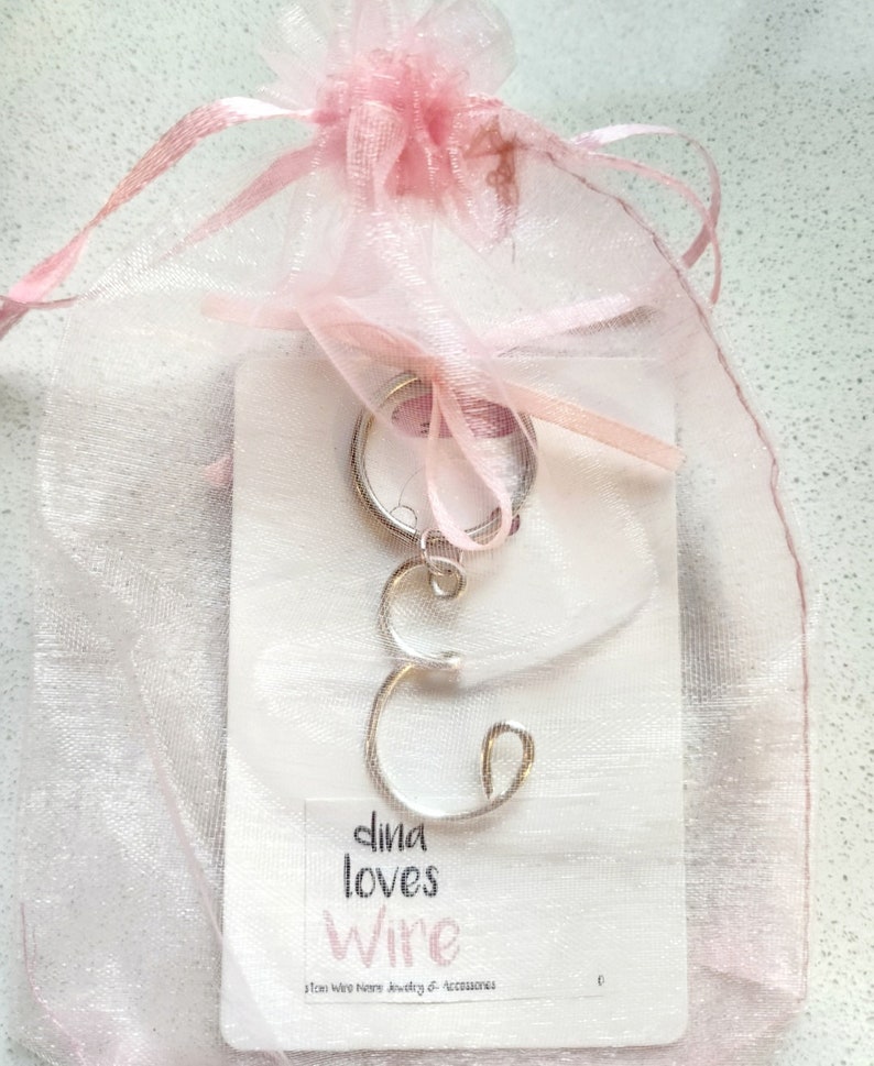 May include: A silver wire letter 'C' charm on a key ring, packaged in a pink organza bag. The card reads "dina loves wire" and "Dina Wire Home Jewelry & Accessories".