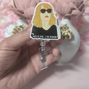 May include: A retractable badge reel featuring a cartoon illustration of a woman with sunglasses and the text "HELP ME, I'M POOR." The reel has a clear plastic body and a metal clip.