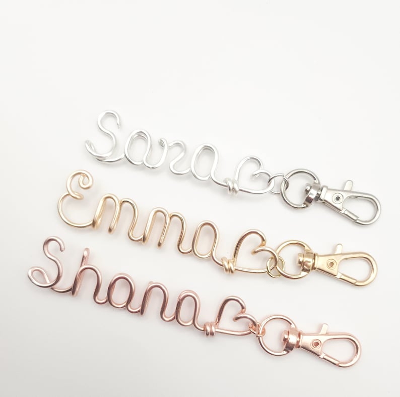 May include: Three personalized keychains with names spelled out in wire and a heart charm. The keychains are silver, gold, and rose gold.