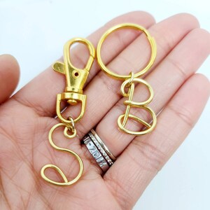 May include: Gold-colored metal keychains with the letters "S" and "B". The keychains have a lobster clasp and a keyring. The letters are in a cursive font.