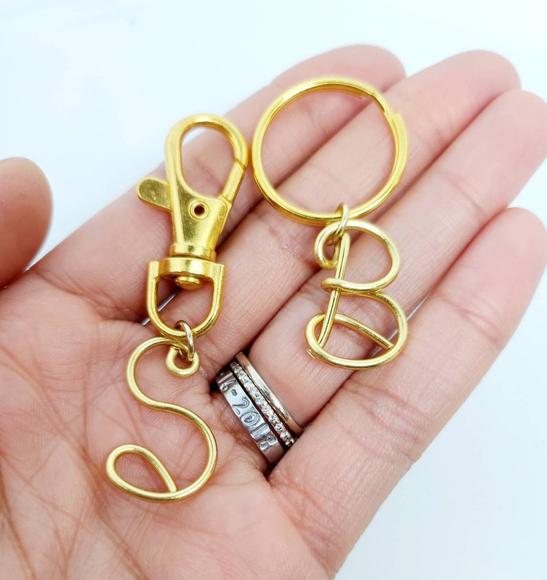 May include: Two gold keychains with the letters "S" and "B" made of wire. The keychains are attached to a gold key ring and a gold clasp.
