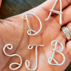 May include: Five silver wire letters, A, D, S, J, and B, are displayed on a hand. The letters are made of wire and are shaped like cursive letters.