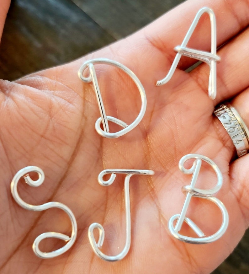 May include: Five silver wire letters, A, D, S, J, and B, are displayed on a hand. The letters are made of wire and are shaped like cursive letters.