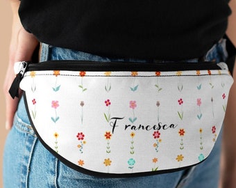Op de afbeelding: Een witte heuptas met een bloemenpatroon en de naam "Francesca" erop gedrukt. De heuptas wordt gedragen om de taille van een persoon die een zwart shirt en een blauwe jeans draagt.