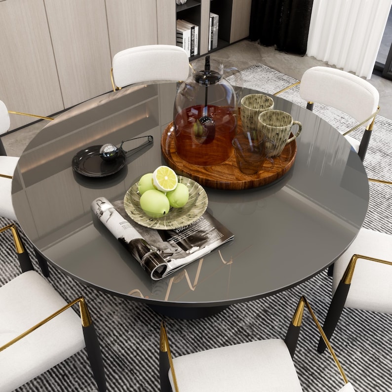 May include: A round glass table with a black top and gold accents. The table is set with a magazine, a plate of lemons, a wooden tray with two mugs and a glass cloche.