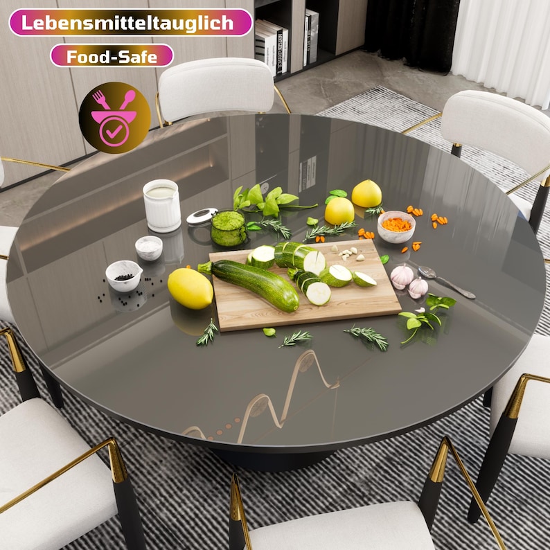 May include: A round, dark gray table with a glass top. The table is set with a wooden cutting board, zucchini, lemons, garlic, and herbs. The table is surrounded by white chairs with gold accents. The table is labeled "Lebensmitteltauglich Food-Safe".