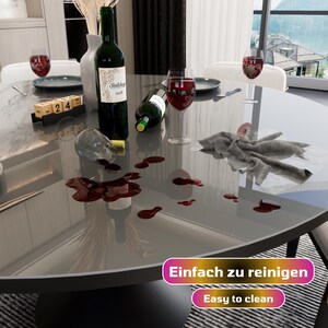 May include: A gray round table with a glass top. Red wine has been spilled on the table, and a cloth napkin is lying on the table. The text "Einfach zu reinigen" and "Easy to clean" is visible on the table.
