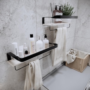 May include: Two black metal shelves with wooden shelves, mounted on a white tiled wall. The top shelf has a small potted plant, two bottles of liquid, and a white towel. The bottom shelf has a variety of bottles and tubes of toiletries, and a white towel hanging from the shelf.