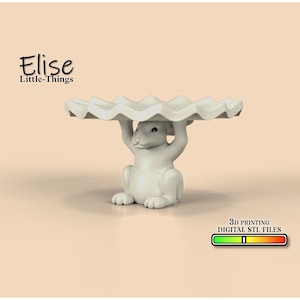 Peut inclure: Un support à gâteau en céramique blanche avec un bord ondulé, soutenu par une figurine de lapin blanc. Le support est conçu pour l'impression 3D et est disponible en fichier STL numérique.
