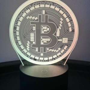 Puede incluir: Un logotipo de Bitcoin blanco impreso en 3D con el texto "Bitcoin Digital Decentralized Peer to Peer" alrededor del borde, iluminado por una base de luz LED multicolor.