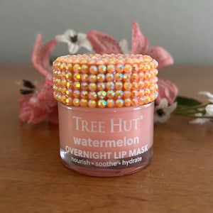 Puede incluir: Un peque&ntilde;o frasco transparente de mascarilla labial nocturna de sand&iacute;a Tree Hut con crema rosa claro en su interior. La tapa est&aacute; cubierta con peque&ntilde;os diamantes de imitaci&oacute;n de color naranja iridiscente. El texto de la etiqueta dice: Tree Hut watermelon OVERNIGHT LIP MASK.