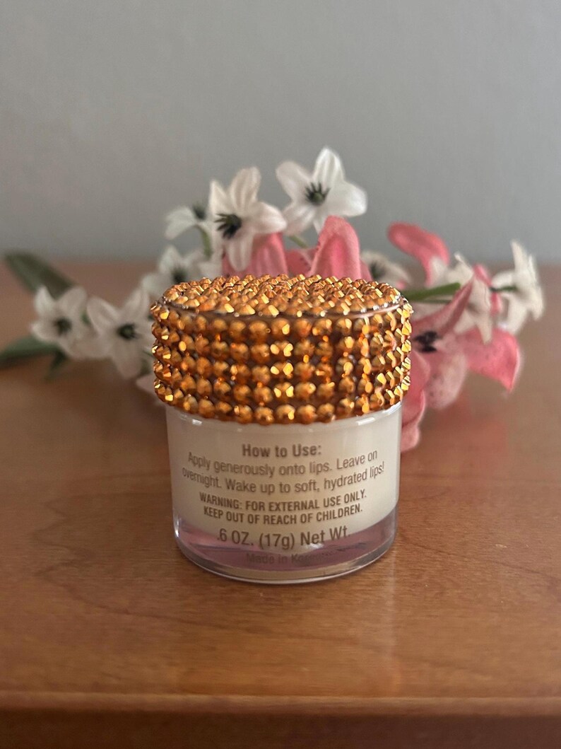 May include: A small, clear cylindrical jar of lip treatment with a lid covered in sparkling gold-toned rhinestones. The label reads: How to Use: Apply generously onto lips. Leave on overnight. Wake up to soft, hydrated lips! WARNING: FOR EXTERNAL USE ONLY. KEEP OUT OF REACH OF CHILDREN. .6 OZ. (17g) Net Wt.