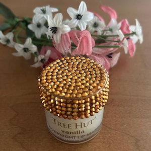 May include: A small cylindrical jar of Tree Hut vanilla overnight lip mask with a lid covered in sparkling gold rhinestones. The white label features black text reading Tree Hut, vanilla, and overnight lip mask, with small white and pink flowers.