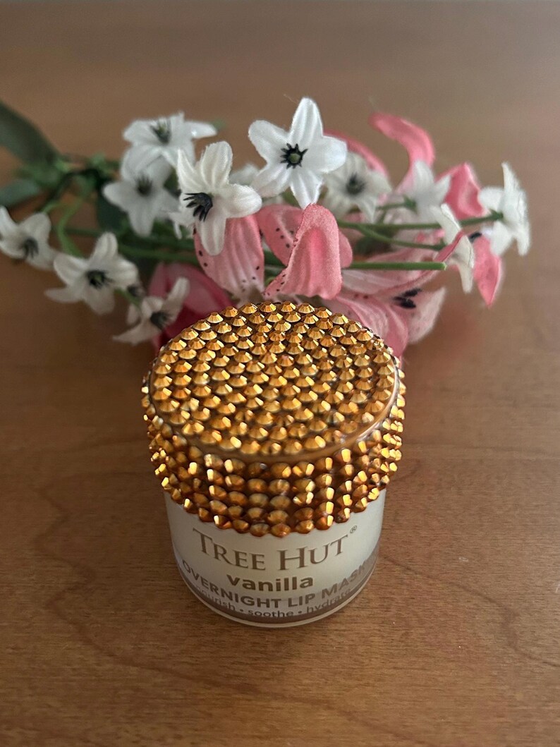 May include: A small cylindrical jar of Tree Hut vanilla overnight lip mask with a lid covered in sparkling gold rhinestones. The white label features black text reading Tree Hut, vanilla, and overnight lip mask, with small white and pink flowers.