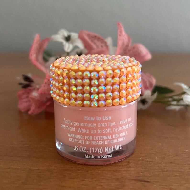 Puede incluir: Un peque&ntilde;o frasco transparente de tratamiento labial color melocot&oacute;n con una tapa cubierta de pedrer&iacute;a naranja iridiscente. La etiqueta dice: How to Use: Apply generously onto lips. Leave on overnight. Wake up to soft, hydrated lips! 17g Net Wt.