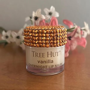 May include: A small, cylindrical jar of Tree Hut vanilla overnight lip mask. The lid is covered in numerous small, reflective, amber-colored rhinestones. The clear plastic container features white text on the front label reading Tree Hut vanilla overnight lip mask.