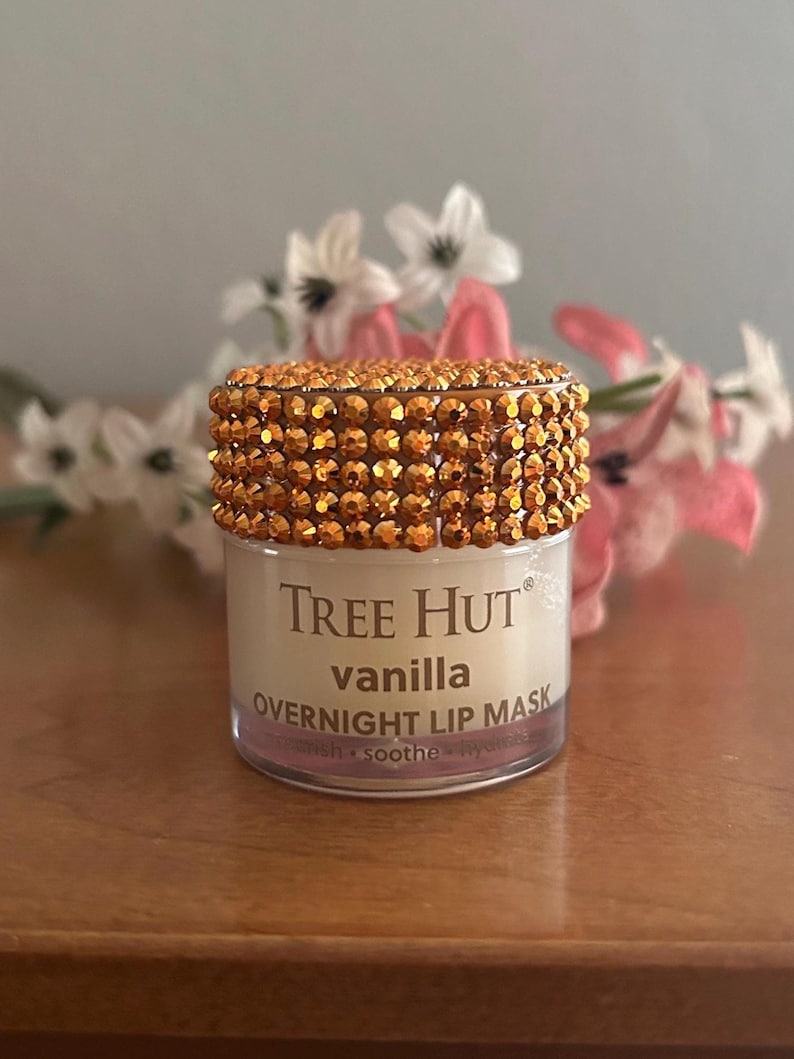 May include: A small, cylindrical jar of Tree Hut vanilla overnight lip mask. The lid is covered in numerous small, reflective, amber-colored rhinestones. The clear plastic container features white text on the front label reading Tree Hut vanilla overnight lip mask.