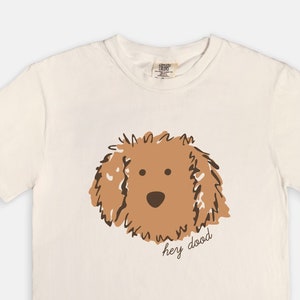 May include: Cream-coloured t-shirt with a brown doodle dog illustration. The dog's face is centered, with the phrase "hey dood" in a handwritten font below. The shirt has short sleeves and a crew neck.