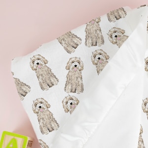 May include: A white baby blanket with a soft, plush texture. The blanket features a repeating pattern of brown and white doodle-style dogs. The blanket is folded in half and has a ruffled edge.