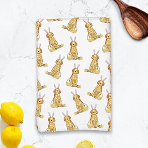 May include: A white kitchen towel with a repeating pattern of golden doodle dogs wearing bunny ears and holding baskets of Easter eggs.