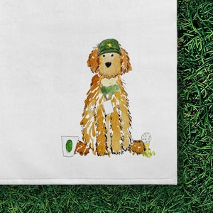 May include: A white kitchen towel featuring a watercolor illustration of a golden-colored dog wearing a green golf cap and a green neck scarf with a white tag. To the left is a white cup with a green circle, and to the right is a golf ball on a tee.