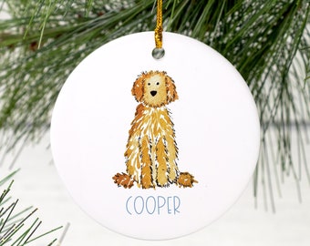 May include: A round, white ceramic ornament with a watercolor illustration of a golden doodle dog. The dog is sitting and the ornament has the name "COOPER" written in blue lettering.