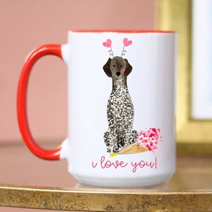 Puede incluir: Taza de cerámica blanca con borde y asa rojos. La taza presenta una ilustración de acuarela de un Braco Alemán de Pelo Corto con corazones rosas sobre su cabeza y un ramo de flores rosas a sus pies. El texto "i love you!" está impreso debajo del perro.