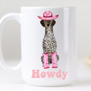 以下が含まれることがあります： ピンクの帽子とピンクのブーツを履いた犬の漫画イラストが描かれた白いセラミック製のコーヒーカップ。犬の下にピンク色の文字で「Howdy」と印刷されています。