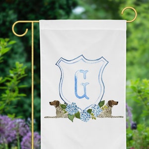 May include: A white garden flag with a blue and white shield featuring the letter "G" in the center. Two brown and white dogs are flanking the shield, and blue and white hydrangeas are below the shield.
