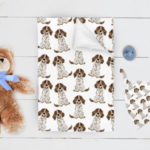Può includere: Coperta per bambini bianca con un motivo ripetuto di cani marroni e bianchi. Sono visibili anche un cappello annodato abbinato e un orsacchiotto marrone con un fiocco blu. Un porta ciuccio blu è sul lato destro dell'immagine.