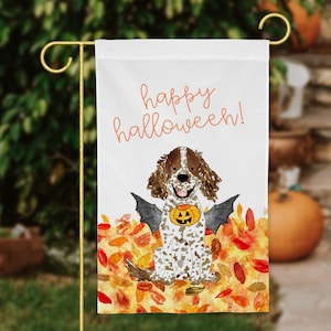 May include: A white garden flag with the text "happy halloween!" in orange. A cartoon dog with brown and white spots is wearing a bat costume and holding a jack-o-lantern. The dog is sitting on a bed of orange, yellow, and red leaves.
