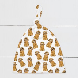 Puede incluir: Un gorro blanco para bebé con un nudo en la parte superior. El gorro tiene un patrón repetido de caniches en miniatura marrones.