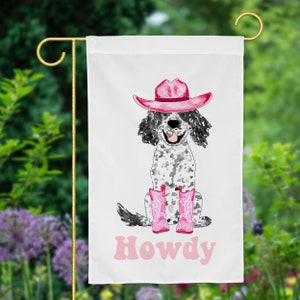 Op de afbeelding: Een witte tuin vlag met een cartoon illustratie van een zwart-witte hond die een roze cowboyhoed en roze laarzen draagt. De hond zit en glimlacht. De tekst "Howdy" is in roze letters onder de hond geschreven.