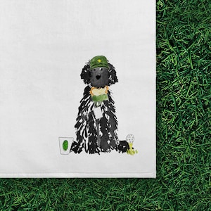 May include: A white tea towel featuring a watercolor illustration of a black curly-haired dog wearing a green golf cap. The dog holds a sandwich, stands next to a white cup with a green logo, and is positioned beside a white golf ball on a yellow tee.