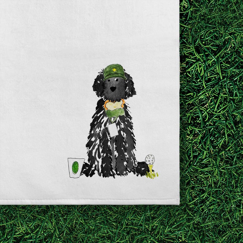 May include: A white tea towel featuring a watercolor illustration of a black curly-haired dog wearing a green golf cap. The dog holds a sandwich, stands next to a white cup with a green logo, and is positioned beside a white golf ball on a yellow tee.