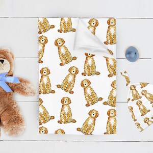 May include: A white baby blanket featuring a repeating pattern of golden-brown doodle dogs, a brown plush teddy bear with a light blue ribbon, a light blue pacifier, and a matching white baby hat with the same golden-brown dog print.