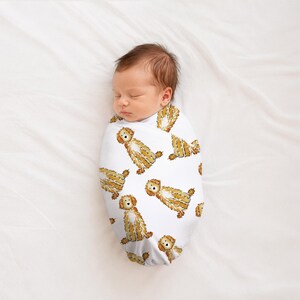 May include: A white swaddle blanket featuring a repeating pattern of illustrated golden-brown dogs with shaggy fur. The fabric is wrapped snugly around a sleeping infant, displaying the canine print clearly across the entire surface of the wrap.