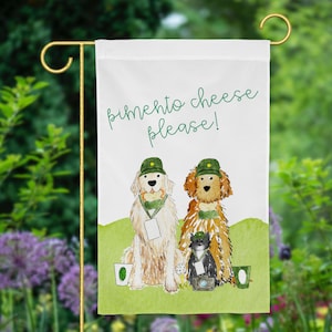 May include: A white garden flag with a green border features two dogs wearing green hats and holding a bone. The text "pimento cheese please!" is written in green cursive.