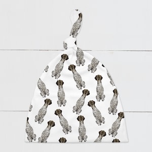 May include: White baby hat with a knot on top. The hat has a repeating pattern of brown and white spotted dogs.