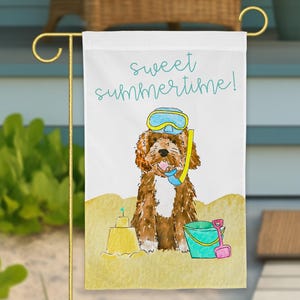 May include: A white garden flag featuring a brown doodle dog wearing a yellow and blue snorkel mask on a sandy beach. The text sweet summertime! is printed in teal above the dog. A yellow sandcastle and a teal bucket with a pink shovel are visible.