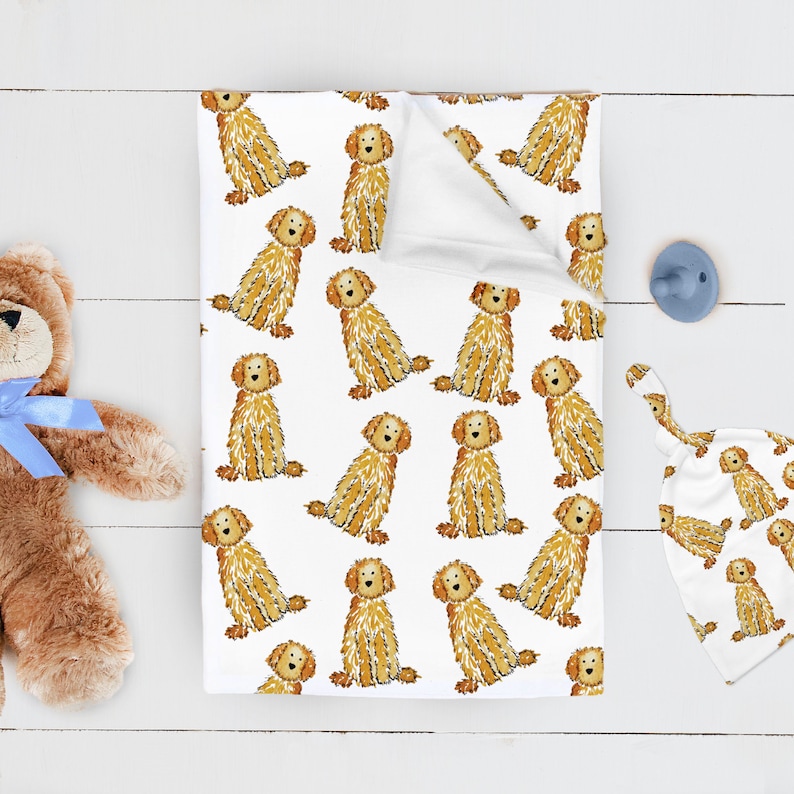 May include: A white baby blanket featuring a repeating pattern of golden-brown doodle dogs, accompanied by a matching white baby hat with the same dog print, a blue pacifier, and a brown plush teddy bear with a light blue ribbon around its neck.