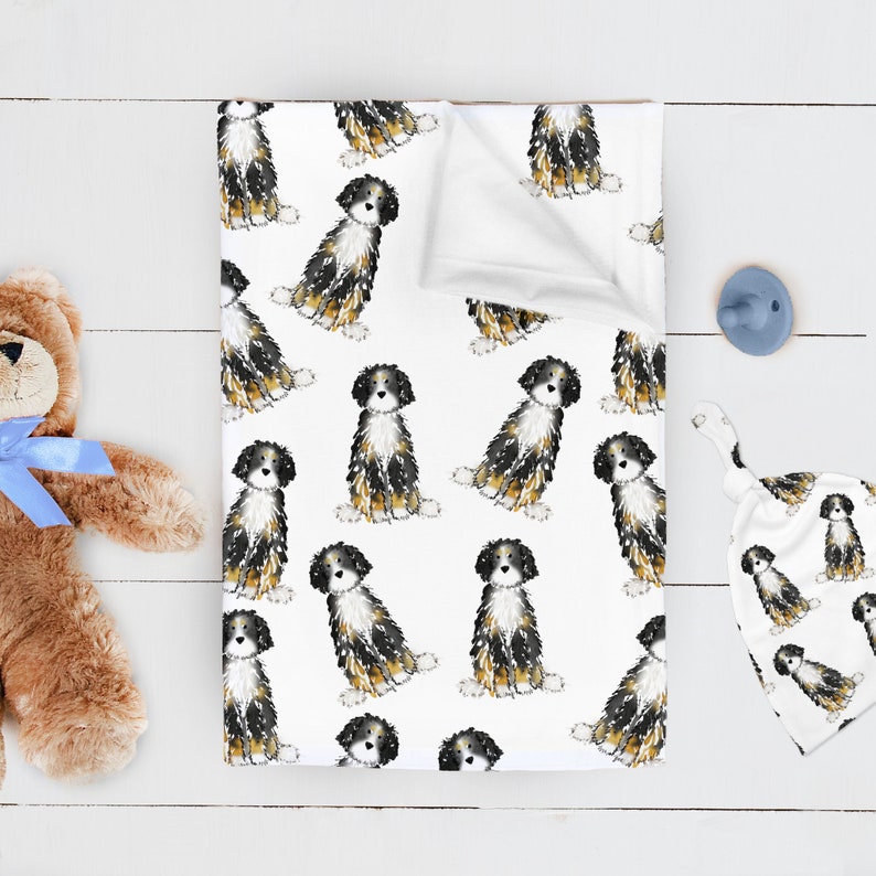 May include: A white baby blanket featuring a repeating watercolor print of tricolor Bernese Mountain Dog puppies in black, white, and tan. The blanket is folded at the top corner, revealing a plain white underside, and has a soft fabric texture.