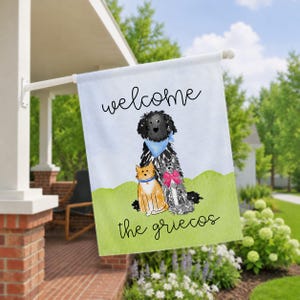 May include: A rectangular garden flag featuring a watercolor illustration of a black dog with a blue bandana, a grey dog with a pink bow, and an orange cat. The text reads welcome at the top and the griecos at the bottom on a green and blue background.
