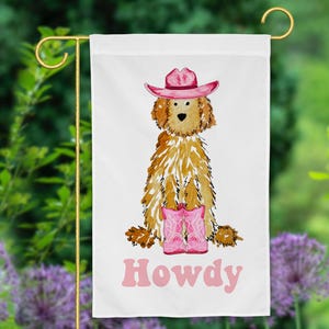 Puede incluir: Un perro doodle marrón y blanco con un sombrero de vaquero rosa y botas de vaquero rosas. El perro está sentado y el texto "Howdy" está escrito debajo del perro en rosa.