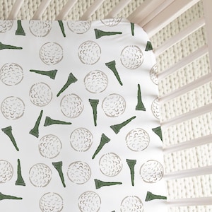 May include: White crib sheet with a repeating pattern of green golf tees and brown golf balls.