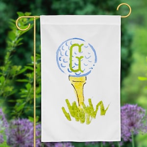 Op de afbeelding: Een witte tuin vlag met een blauwe en groene golfbal met de letter "G" erop. De golfbal staat op een gele tee en er is groen gras aan de onderkant van de vlag.