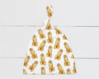 May include: White baby hat with a knot on top. The hat has a repeating pattern of golden retriever dogs.