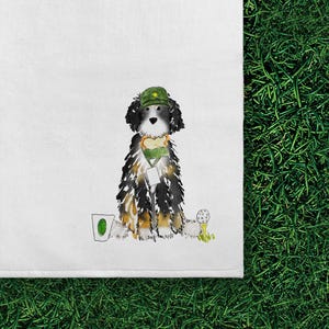May include: A white tea towel featuring a watercolor illustration of a black, white, and tan dog wearing a green baseball cap. The dog has a sandwich around its neck, a white tag, a small golf ball on a tee, and a green drink cup nearby.